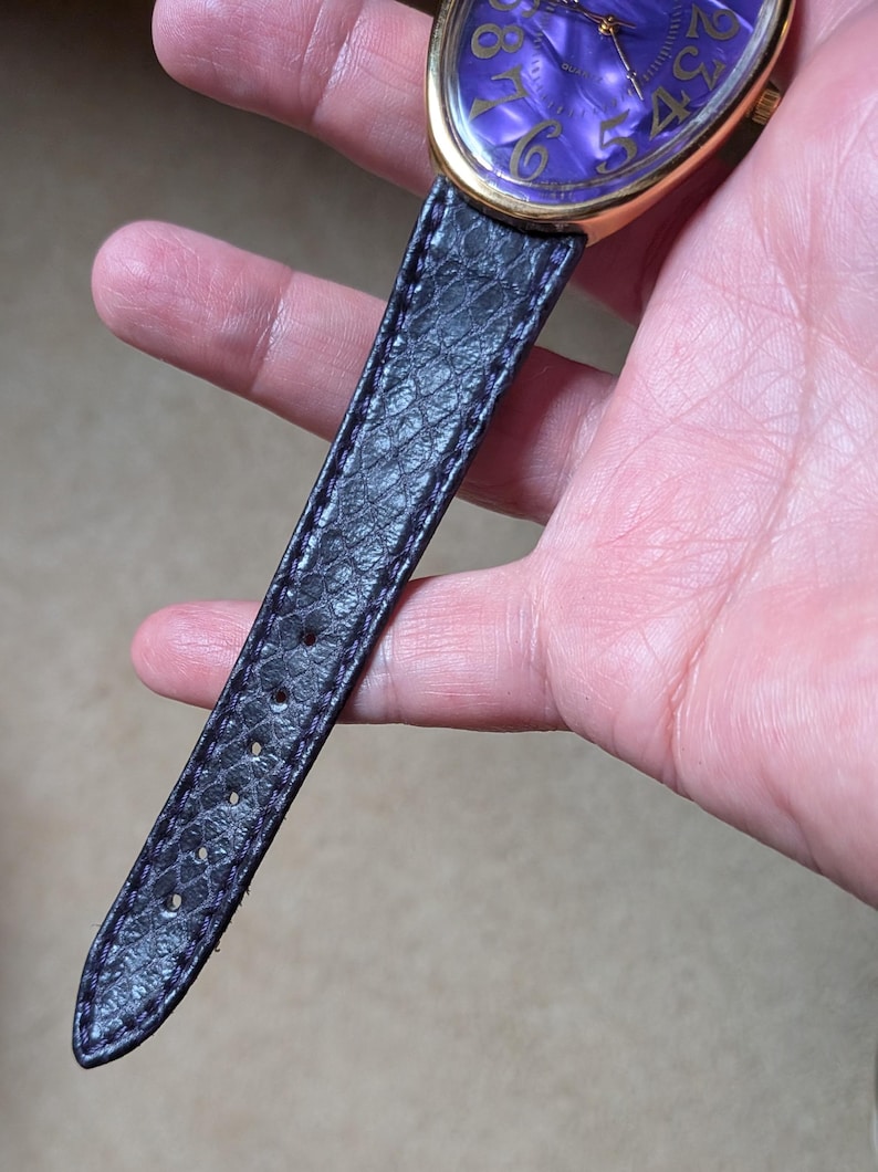 May include: A gold-toned wristwatch with a purple face and black leather band. The watch face has gold numerals and a swirling pattern. The black leather band has a textured, snake-like pattern.