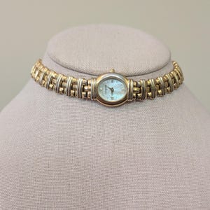 May include: A gold and silver-toned watch necklace with an oval watch face. The watch face has a light blue iridescent finish and Roman numeral hour markers. The watch band is made of rectangular links.