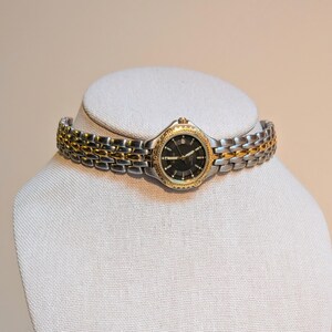 May include: A two-tone wristwatch with a black dial and gold-tone accents. The watch features a silver and gold band. The watch is displayed on a white mannequin head.
