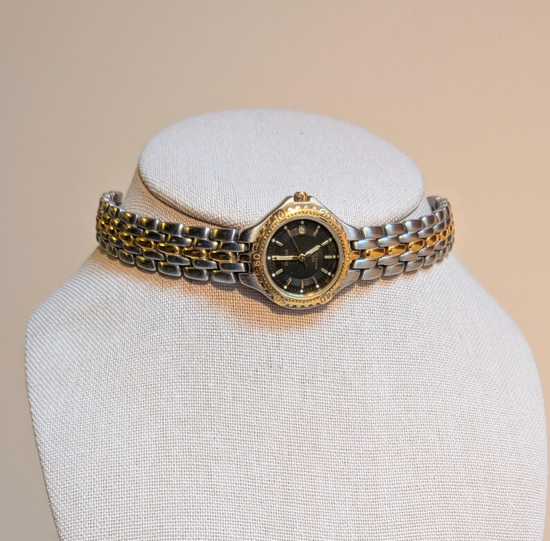 May include: A two-tone wristwatch with a black dial and gold-tone accents. The watch features a silver and gold band. The watch is displayed on a white mannequin head.