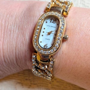 May include: Gold-toned Sofia Vergara quartz watch with a white face and crystal accents. The oval watch face features the number 12 and 6, with the brand name and "Quartz" printed on the face. The watch band is also gold-toned with crystal embellishments.