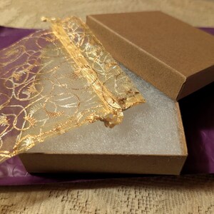 May include: An open brown cardboard box with a matching lid, revealing a white interior. A gold mesh bag with an ornate design is partially inside the box. The box rests on a purple padded envelope.