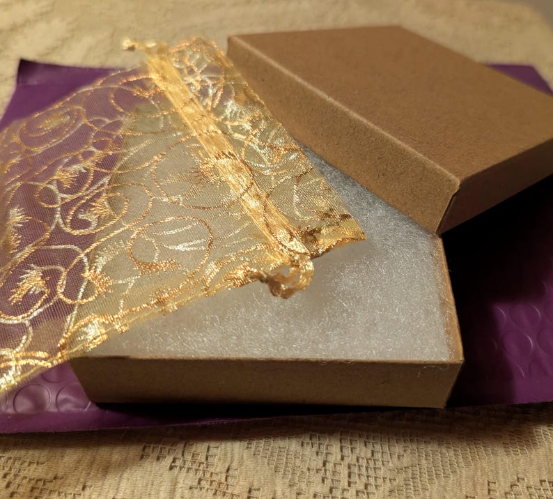 May include: An open brown cardboard box with a matching lid, revealing a white interior. A gold mesh bag with an ornate design is partially inside the box. The box rests on a purple padded envelope.