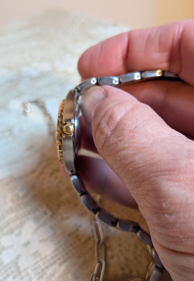 May include: A two-tone wristwatch with a silver-colored band and a gold-colored bezel. The watch face is partially visible, showing gold accents. The watch is held by a hand, showcasing the side profile and the band's links.