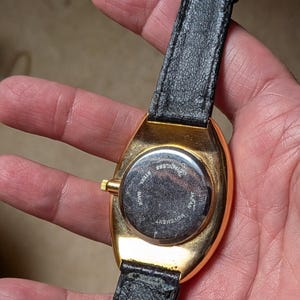 May include: A gold-toned wristwatch with a black leather band. The watch has a rectangular face and a stainless steel back. The band is secured with a gold-colored buckle. The watch is held in a person's hand.