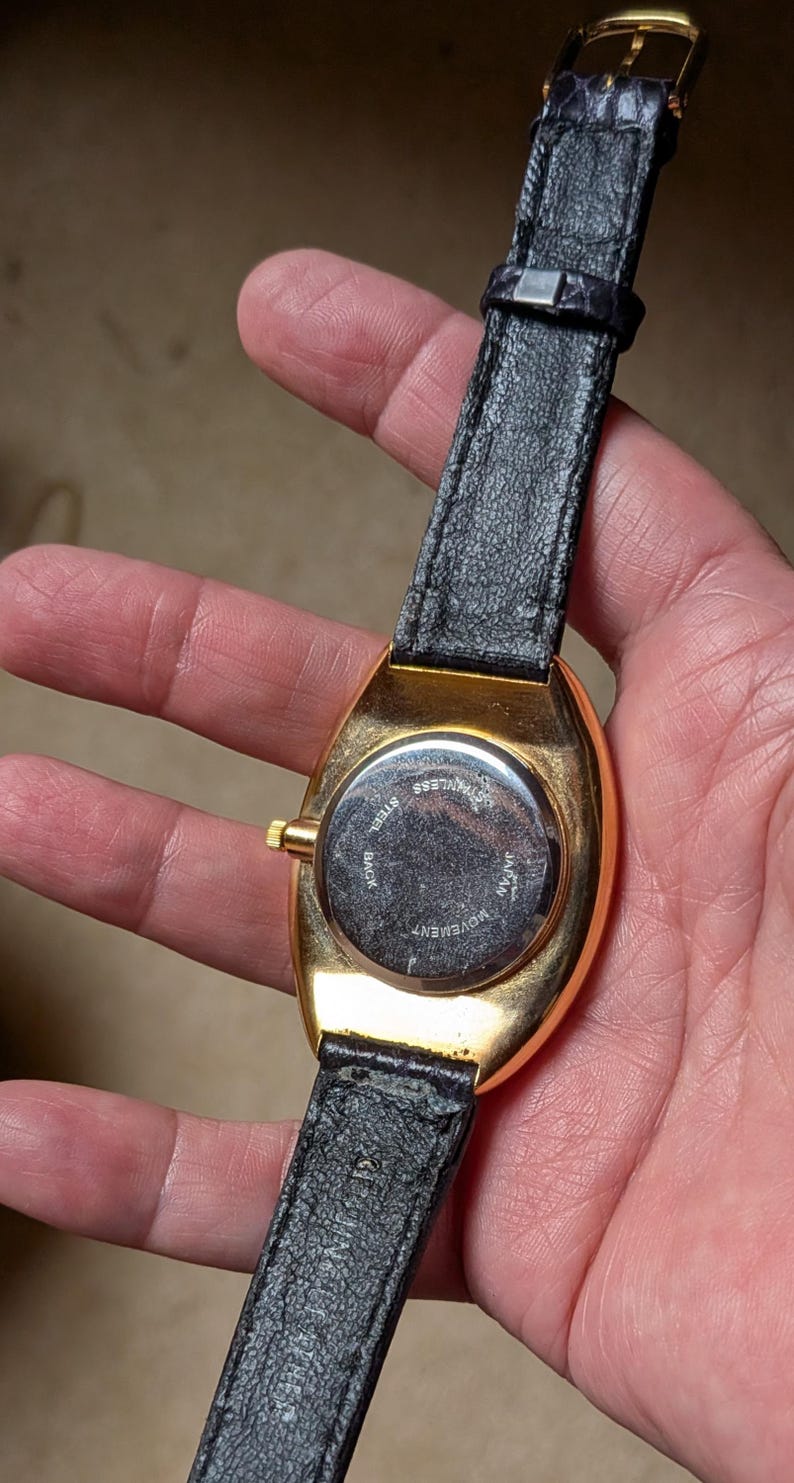 May include: A gold-toned wristwatch with a black leather band. The watch has a rectangular face and a stainless steel back. The band is secured with a gold-colored buckle. The watch is held in a person's hand.