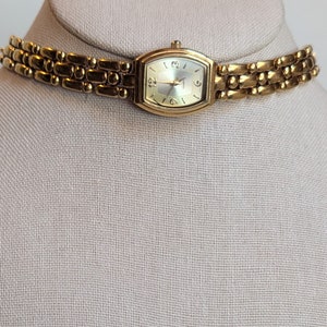 Puede incluir: Un collar de reloj dorado con una esfera rectangular. La esfera tiene un fondo claro con números y manecillas negras. La correa del reloj está hecha de eslabones rectangulares conectados.