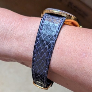 May include: A gold-toned wristwatch with a dark blue, textured leather band. The band features a scale-like pattern and a gold-colored buckle. The watch face is visible, with a clear crystal and gold-toned bezel.