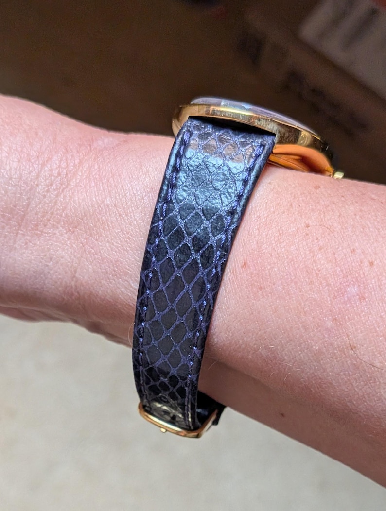 May include: A gold-toned wristwatch with a dark blue, textured leather band. The band features a scale-like pattern and a gold-colored buckle. The watch face is visible, with a clear crystal and gold-toned bezel.