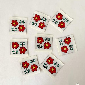 May include: Eight white square fabric labels with a red flower design. Each label features two red flowers with yellow centers and green stems and leaves. The labels are arranged on a white surface.
