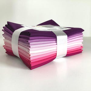 May include: A stack of folded fabric squares in shades of purple and pink, secured with a white ribbon. The colors range from deep violet to light pink, creating a gradient effect. The fabric appears to be cotton or a similar material.