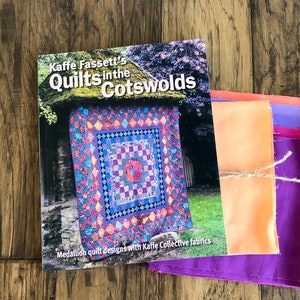 May include: A book titled "Kaffie Fassett's Quilts in the Cotswolds" with a colorful quilt on the cover. The book is next to a stack of fabric swatches in shades of orange, purple, and pink.