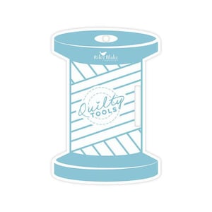 May include: A light blue and white sticker in the shape of a spool of thread. The sticker has the text "Riley Blake Designs" and "Quilty Tools". The spool is a quilting tool.