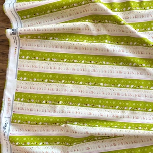 May include: Fabric with a repeating pattern of green and white stripes. The green stripes are interspersed with white stripes featuring small floral and dotted designs. The fabric is folded, showing the edge with text.