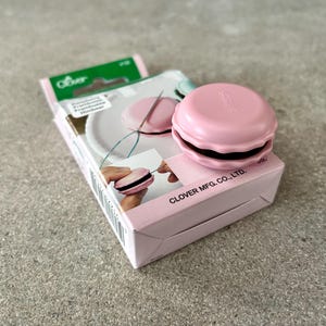 May include: A pink Clover brand needle minder in the shape of a macaron sits atop its packaging. The packaging is pink and white, with images of the product in use. The needle minder is a sewing and embroidery accessory.