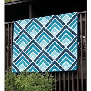 May include: A quilt with a geometric pattern of blue and white chevrons, hanging on a wooden railing. The quilt has a repeating design of dark blue, light blue, and white shapes. The text "MOUNTAIN PEAKS" is at the top.