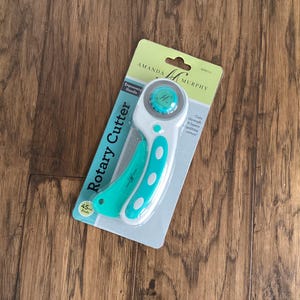 May include: A teal and white rotary cutter by Amanda Murphy, packaged on a light green and white card. The cutter has an ergonomic handle and a 45 mm blade. The packaging reads "Rotary Cutter".