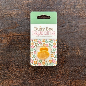 May include: A packaging card for a Busy Bee Thread Cutter. The card is green with a floral pattern and features a yellow bee illustration. The text "Busy Bee Thread Cutter" is printed in black.