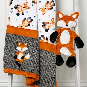 May include: A baby blanket and stuffed fox toy. The blanket features a white background with orange foxes and green leaves. The bottom section is gray with an appliqued fox. The blanket is trimmed with orange fabric. The stuffed fox is orange, white, and black.