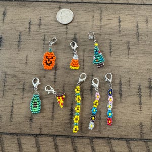 May include: A collection of eight beaded charms with silver-tone lobster clasps, including shapes like a pumpkin, candy corn, Christmas tree, avocado, pizza slice, and three floral strands, shown next to a US dime for scale reference.
