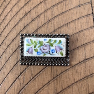 May include: Rectangular silver-toned brooch with a floral design. The brooch features a white background with a painted design of purple, blue, and green flowers and leaves. The edges are framed with a beaded border.