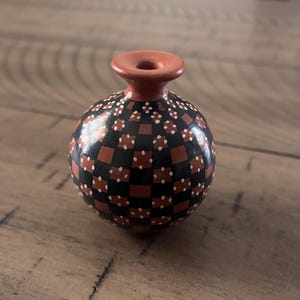 May include: A small, round ceramic vase with a reddish-brown neck and a spherical body. The body is decorated with a checkerboard pattern of black and reddish-brown squares, each with white polka dots. The vase is approximately 4 inches tall.