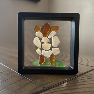 May include: A framed artwork featuring a sea turtle design made from various pieces of sea glass. The sea glass pieces are in shades of brown, white, and green, arranged against a clear background within a black square frame. The artwork is displayed on a wooden surface.