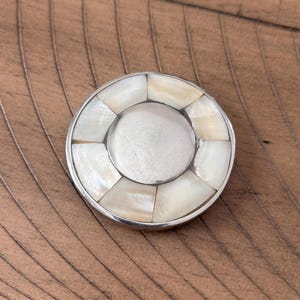 May include: A round, silver-toned object with a central disc and an outer ring of iridescent, rectangular mother-of-pearl segments. The object is set against a brown, textured surface, creating a contrast of cool and warm tones. The design is symmetrical and elegant.