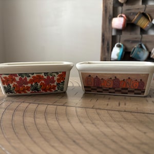 May include: Two rectangular ceramic dishes with cream-colored exteriors. One dish features a design of fall leaves in orange, red, and green. The other dish has the word "Blessings" and pumpkin illustrations. Both sit on a wooden surface.