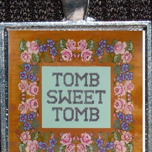 May include: A silver pendant with a brown and gold background featuring a floral border and the text "Tomb Sweet Tomb" in black.