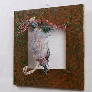 May include: Abstract mixed-media artwork featuring a square, textured, brown frame with a green overlay. Inside the frame, a wire sculpture with red, silver, and blue elements is suspended, creating a three-dimensional effect. The artwork is mounted on a white wall.