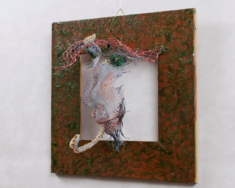 May include: Abstract mixed-media artwork featuring a square, textured, brown frame with a green overlay. Inside the frame, a wire sculpture with red, silver, and blue elements is suspended, creating a three-dimensional effect. The artwork is mounted on a white wall.
