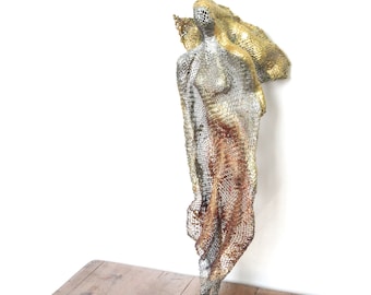 May include: A wire sculpture of a standing figure with a flowing garment. The sculpture is made of silver, gold, and copper-coloured wire. The figure has a mesh-like appearance, with a detailed form and texture. The sculpture is displayed against a white wall.