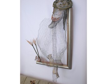 May include: A silver-framed wire sculpture depicting a figure wearing a hat. The sculpture is crafted from fine wire mesh, forming the shape of a person. A small arrangement of flowers is included, adding a delicate touch to the artwork.
