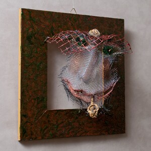 May include: Abstract mixed-media artwork featuring a square, dark brown frame with a textured surface. Inside the frame, a complex arrangement of wire mesh, red netting, and gold accents creates a unique, three-dimensional design. The artwork is suspended by a wire.