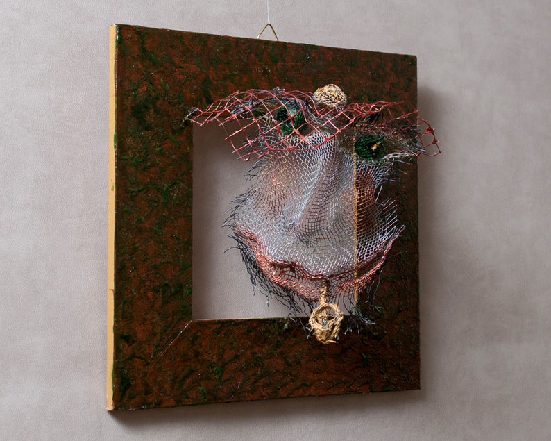 May include: Abstract mixed-media artwork featuring a square, dark brown frame with a textured surface. Inside the frame, a complex arrangement of wire mesh, red netting, and gold accents creates a unique, three-dimensional design. The artwork is suspended by a wire.
