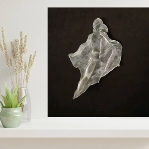 May include: A silver mesh sculpture against a black background. The artwork has a flowing, draped form, resembling a figure or garment. The piece is illuminated, creating highlights and shadows that emphasize its texture and shape. The composition is minimalist, with the sculpture as the focal point.