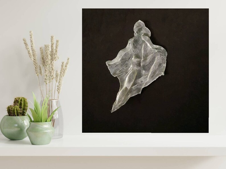 May include: A silver mesh sculpture against a black background. The artwork has a flowing, draped form, resembling a figure or garment. The piece is illuminated, creating highlights and shadows that emphasize its texture and shape. The composition is minimalist, with the sculpture as the focal point.
