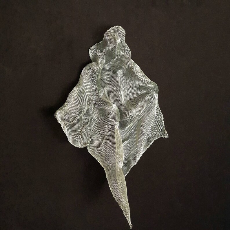 May include: A silver-colored, abstract sculpture made of a woven mesh material. The artwork has a draped, flowing form, with a pointed, elongated section extending downwards. The sculpture is set against a dark brown background.