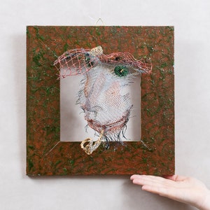 May include: A wire sculpture of a face within a square, textured frame. The face is constructed from wire mesh, with details like a hat and eyes. The frame is a mix of brown and green, creating a unique art piece.