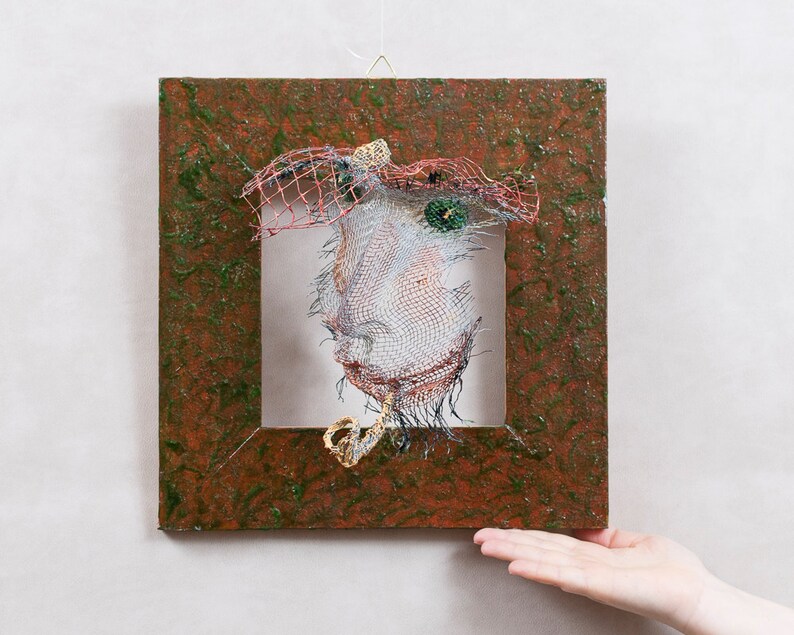 May include: A wire sculpture of a face within a square, textured frame. The face is constructed from wire mesh, with details like a hat and eyes. The frame is a mix of brown and green, creating a unique art piece.
