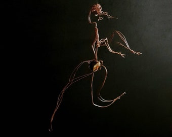 May include: A copper wire sculpture of a figure in motion against a black background. The sculpture has a flowing, abstract design with a central stone element. The figure appears to be mid-stride, with arms outstretched.