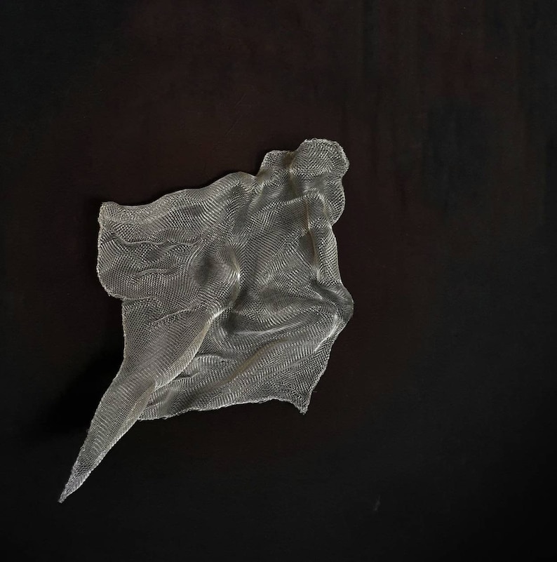 May include: A silver-colored, mesh-like sculpture with a flowing, draped form. The artwork is set against a dark background, creating a contrast that highlights the texture and shape of the piece. The sculpture appears to be made of a flexible, woven material.