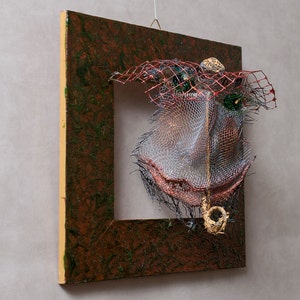 May include: Abstract mixed-media artwork featuring a square wooden frame with a textured brown and green surface. The frame holds a complex arrangement of wire mesh, red netting, and metallic accents, creating a three-dimensional effect. A small, gold-colored element hangs from the bottom.
