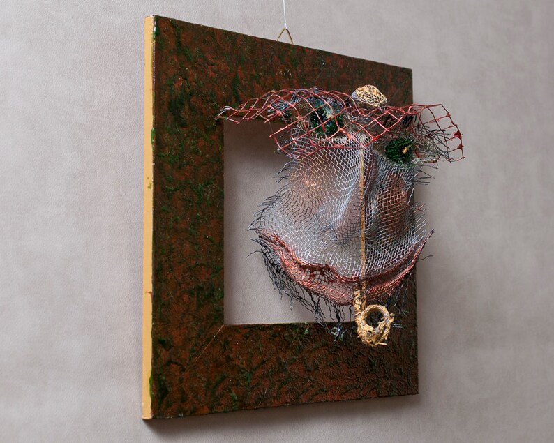 May include: Abstract mixed-media artwork featuring a square wooden frame with a textured brown and green surface. The frame holds a complex arrangement of wire mesh, red netting, and metallic accents, creating a three-dimensional effect. A small, gold-colored element hangs from the bottom.