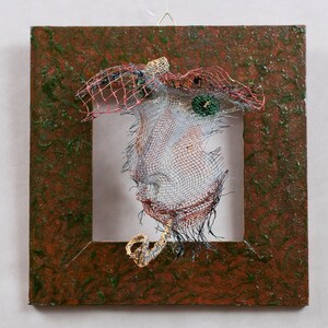 May include: A mixed-media artwork featuring a wire mesh face within a square frame. The face is constructed from wire, with a red and black mesh headdress. The frame has a textured brown and green surface, creating a unique artistic piece.