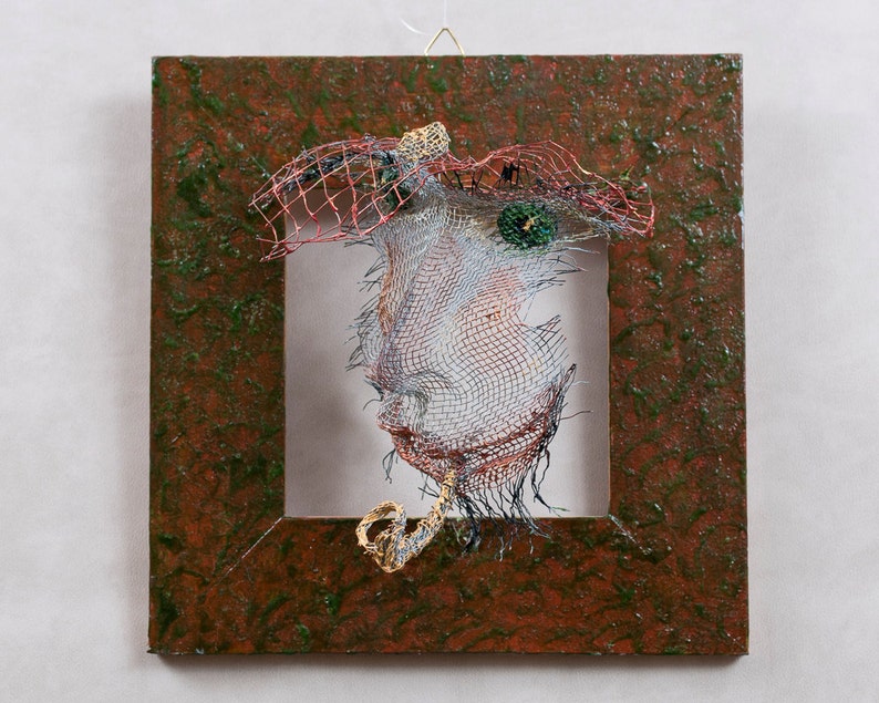 May include: A mixed-media artwork featuring a wire mesh face within a square frame. The face is constructed from wire, with a red and black mesh headdress. The frame has a textured brown and green surface, creating a unique artistic piece.