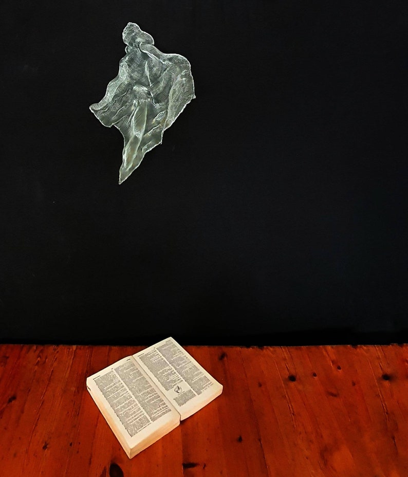 May include: An open book with text lies on a red-brown wooden floor. Above, a translucent, abstract sculpture hangs against a black background. The sculpture appears to be a figure or form.