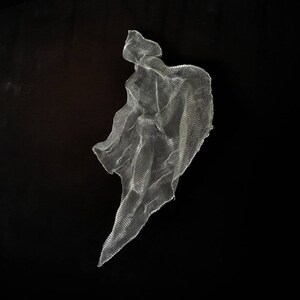 May include: A silver-colored, abstract sculpture made of a fine mesh material. The artwork has a flowing, draped form against a black background, creating a contrast of light and shadow. The sculpture appears to be suspended.