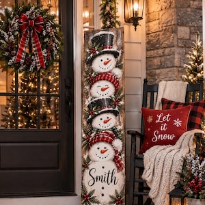 May include: A tall wooden porch sign featuring a vertical stack of four snowmen with hats and scarves, with the name Smith printed at the bottom. The sign is displayed on a porch decorated with a wreath, a welcome mat, lanterns, and greenery.